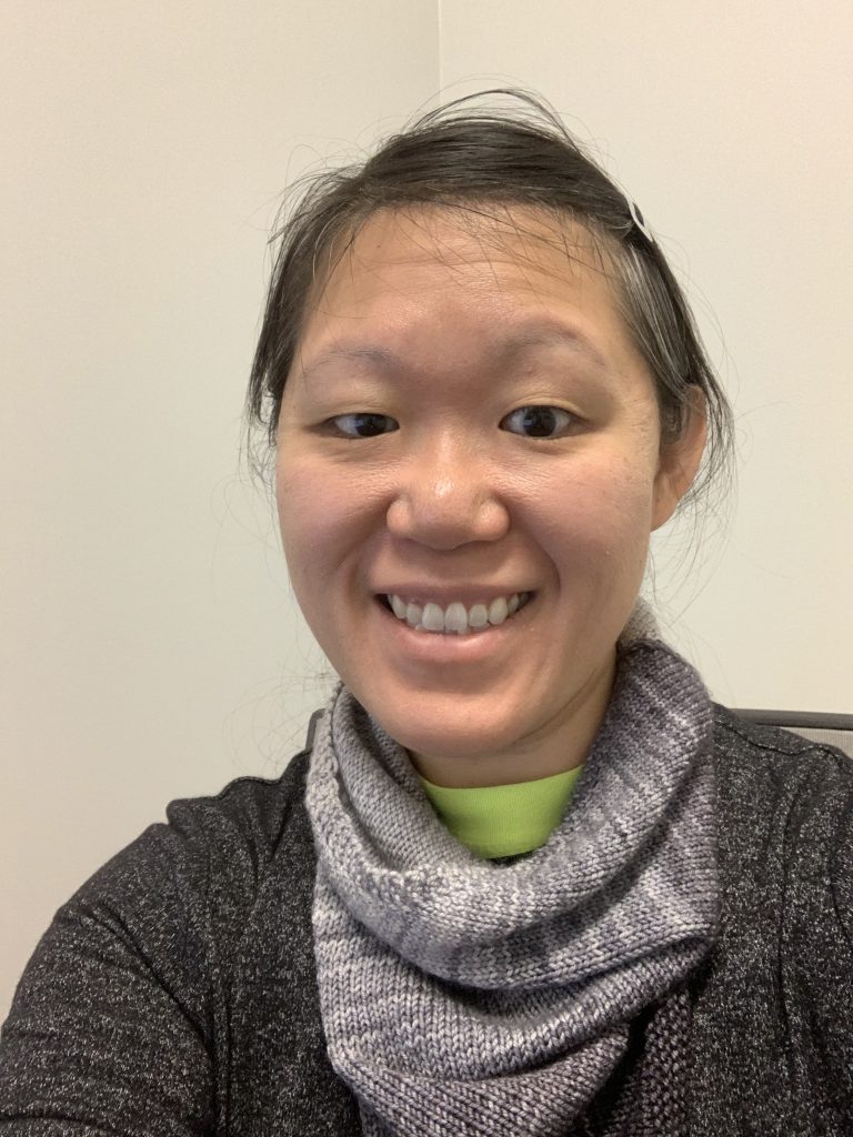 East Asian adult woman smiling, wearing neckwarmer and sweater.