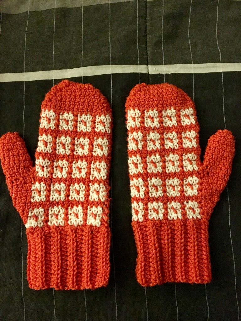 Orange mittens with white squares laying on a black, white, and gray striped comforter.